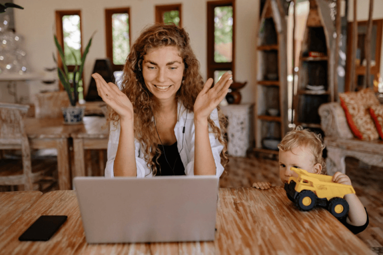 Professional woman with curly hair gesturing during video call while toddler plays with toy truck nearby, showing confident parent managing work-family balance