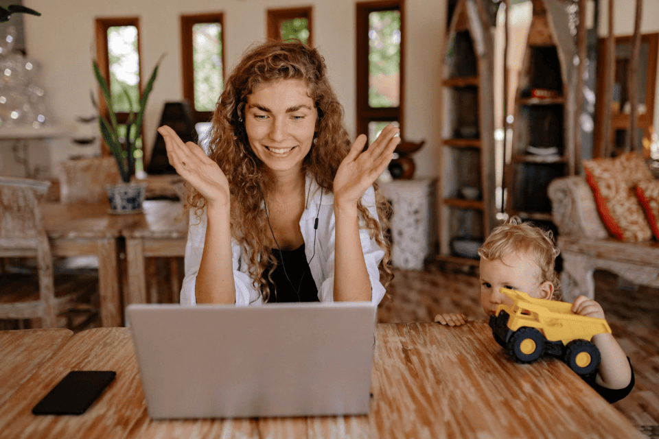 Professional woman with curly hair gesturing during video call while toddler plays with toy truck nearby, showing confident parent managing work-family balance